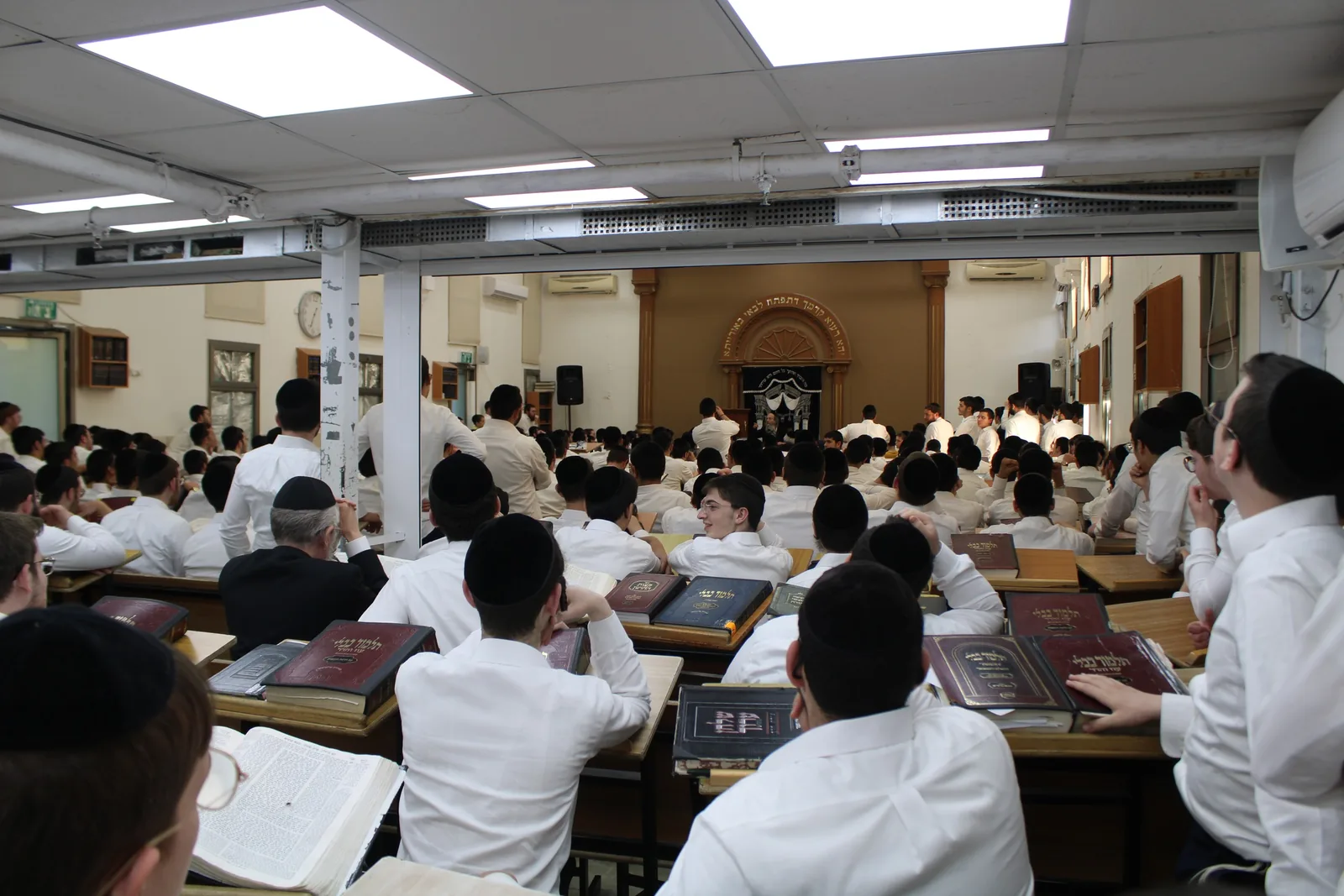 מבט מהספסלים האחוריים על בית המדרש המלא בשעת לימוד, גמרות פתוחות על הסטנדרים